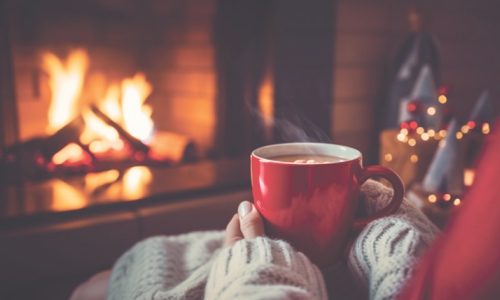A woman holds a hot cocoa while sitting in front of the fireplace