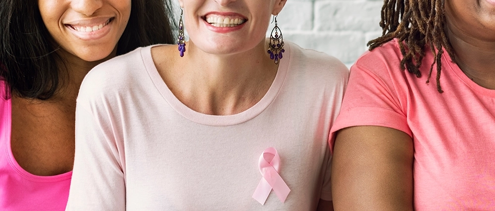 Three women sit and smile while wearing pink shirts and breast cancer ribbons.
